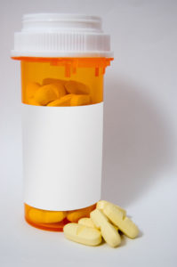A standard orange prescription bottle full of yellow pills. The information on the label has been covered. A few pills sit outside the bottle, at its base.